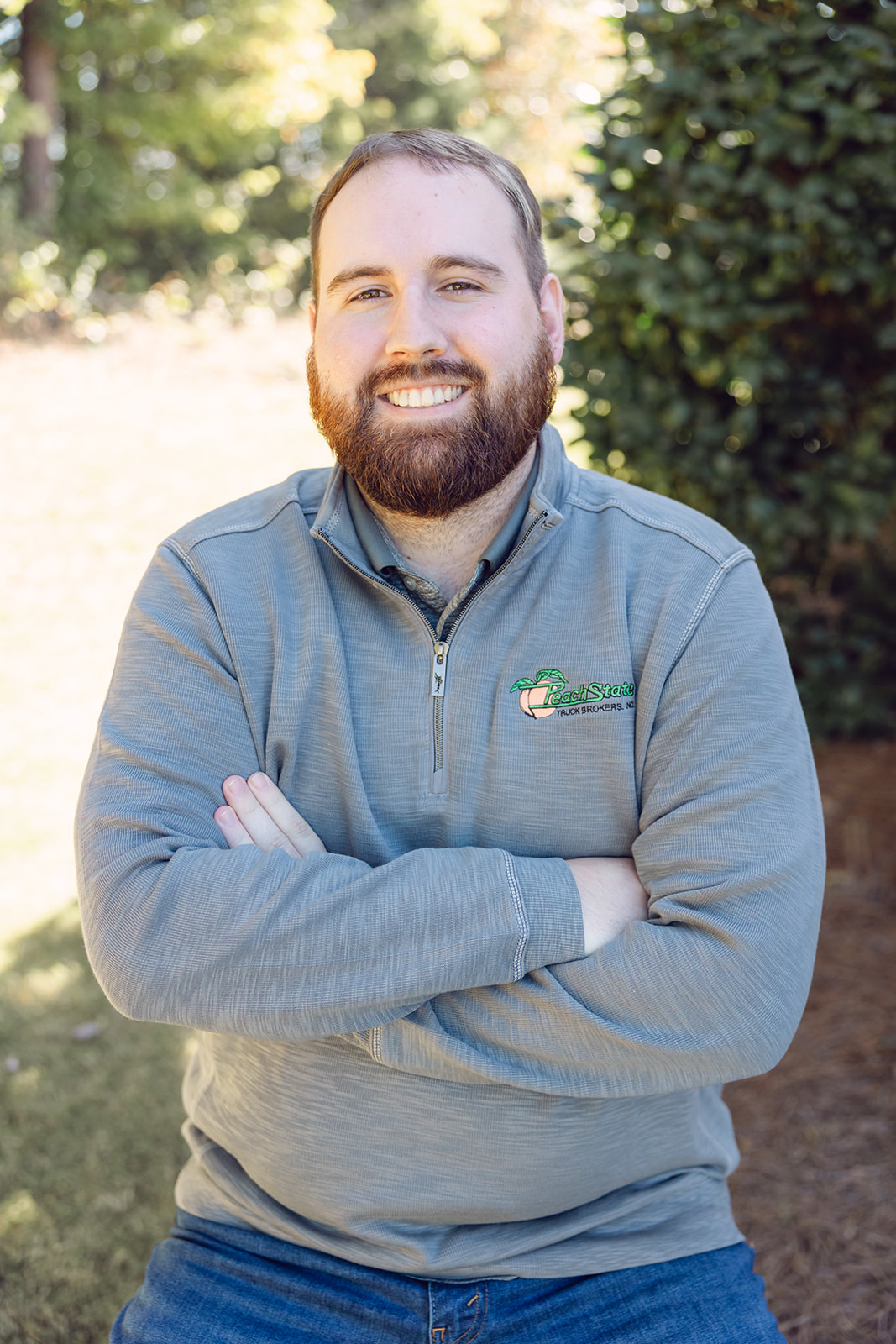 A man with beard and arms crossed in front of bushes.